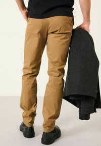 Beige Baumwollhosen mit schmaler Passform, kombiniert mit schwarzen Schuhen. Die Person hält eine dunkelgraue, strukturierte Jacke über ihrem Arm.