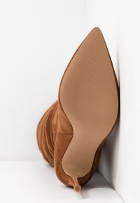 Botte stiletto en daim marron à bout pointu reposant contre un coin blanc, montrant la semelle propre et la fine pointe du talon.