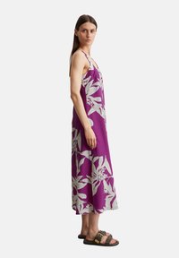 Paarse maxi-jurk met wit bloemenpatroon, mouwloos ontwerp, vloeiende stof en een zijsplit; gecombineerd met bruine sandalen.