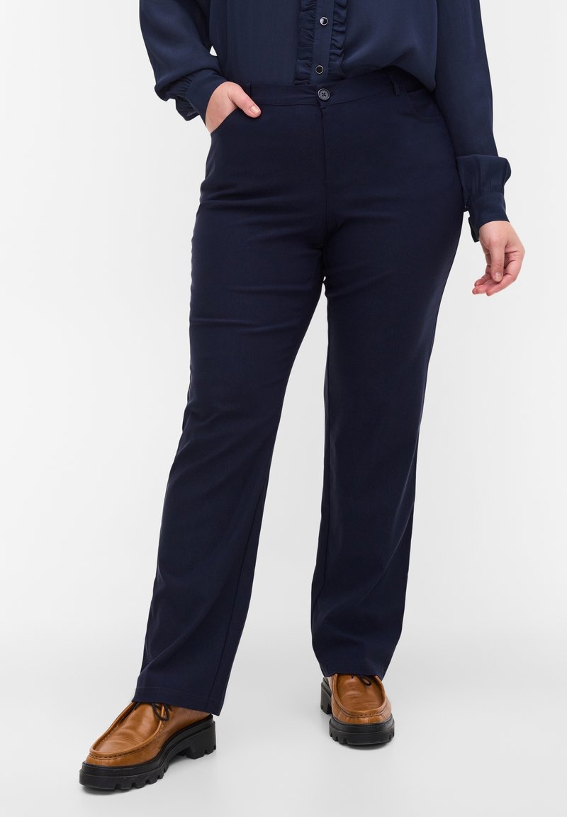 Femme portant un pantalon bleu marine et un chemisier assorti avec une patte de boutonnage à volants, des chaussures à lacets marron à semelle épaisse, une main dans la poche.
