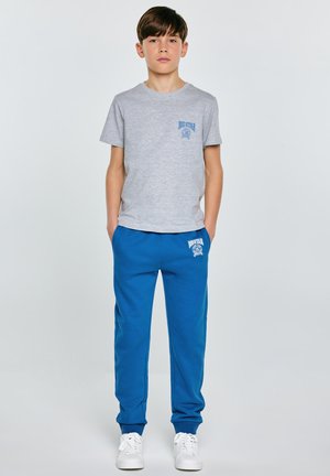 Chlapec stojící s rukama v kapsách, oblečený v šedém tričku Big Star, modrých teplácích a bílých teniskách na jednobarevném pozadí.