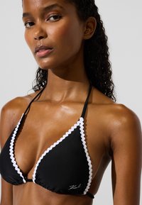 Top bikini nero a forma triangolare, con bordi ondulati bianchi e spalline regolabili. Presenta un logo distintivo alla base.