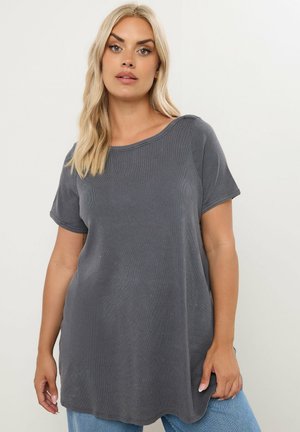 Kobieta o długich blond włosach, ubrana w luźną ciemnoszarą koszulkę z krótkim rękawem i jasnoniebieskie jeansy na jednolitym tle.