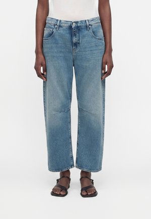 Person iført lyseblå rette cropped jeans og sorte åbne sandaletter, stående mod en hvid baggrund.