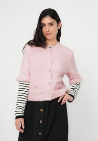 Ροζ υφασμάτινη ζακέτα με κοντά μανίκια, κλείσιμο με κουμπιά και δύο μπροστινές τσέπες. Φορεμένη πάνω από μαύρη φούστα με καφέ κουμπιά και ριγέ μανίκια.