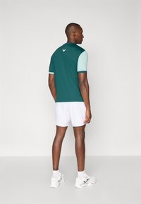 Mørkegrønn kortermet treningsskjorte med lyseblå ermer, hvite shorts og joggesko, sett fra baksiden. Logo på nakken.