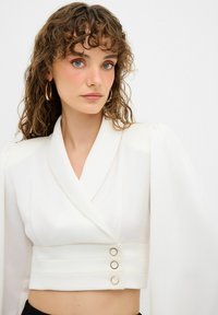 Blusa blanca corta con diseño de envoltura, mangas largas abullonadas y tres botones decorativos dorados en la cintura. Textura suave.
