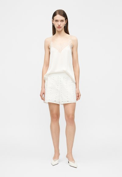 Kobieta ubrana w białą koszulkę na ramiączkach z koronkową lamówką, białe krótkie spodenki z ażurowym wzorem oraz białe szpilki z ostrym noskiem, stojąca na jednolitym tle.