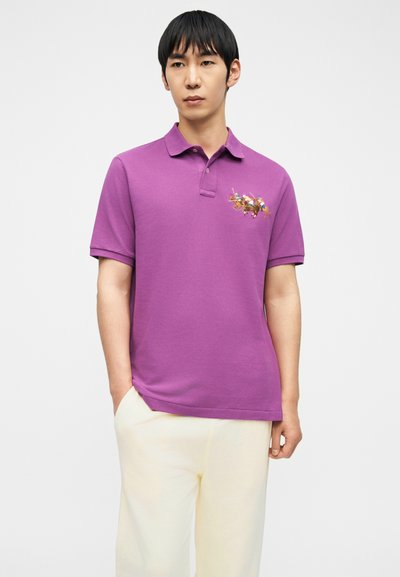 Άνδρας που φοράει μωβ πόλο μπλούζα με γραφικό παίκτη πόλο με άλογο στο στήθος και ανοιχτό μπεζ παντελόνι, στέκεται μπροστά σε απλό φόντο.