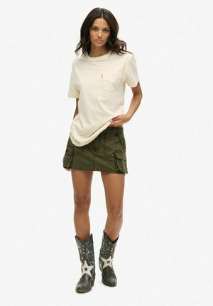 Beige Baumwoll-T-Shirt mit einer Brusttasche, kombiniert mit einem olivgrünen Cargo-Rock mit aufgesetzten Taschen und schwarzen Cowboy-Stiefeln mit Sternen.