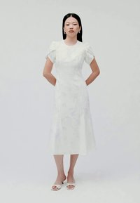 Weißes Midi-Kleid mit strukturierten Blumenmustern, kurzen Puffärmeln und tailliertem Oberteil. Getragen mit weißen Sandalen mit offenen Zehen.