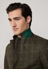 Zielono-brązowy płaszcz w desenie houndstooth z detalem kołnierza w jednolitym zielonym kolorze. Projekt cechuje nowoczesny krój i teksturowana tkanina z delikatnym wzorem w kratę.