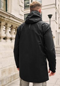 Une veste imperméable noire avec une capuche, présentant une texture lisse, une longueur longue et des accents réfléchissants à l'arrière.