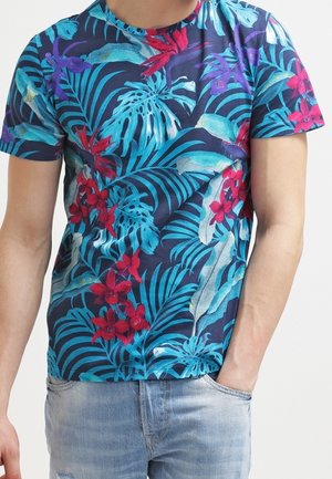 Man wearing a short-sleeve navy shirt with bright blue leaves and red floral patterns, paired with light blue faded jeans.