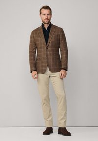 Brauner kariert Blazer mit tailliertem Schnitt, Ein-Knopf-Verschluss, kombiniert mit einem schwarzen Hemd mit Kragen und beigen Hosen. Braune Stiefeletten.