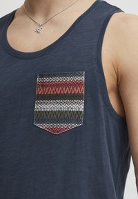 Tricou fără mâneci de culoare bleumarin, cu un buzunar modelat ce prezintă un design geometric în roșu, verde și alb, realizat dintr-un material ușor, cu o textură netedă.