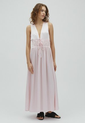 Fiatal nő göndör hajjal, világos rózsaszín ujjatlan maxiruha mély V-kivágással és fekete szandállal, sima háttér előtt áll.