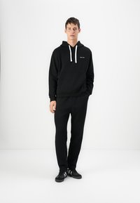 Black hoodie with white drawstrings, kangaroo pocket, and logo on the chest. Paired with black pants and black sneakers.