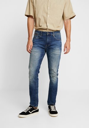 Mand iført falmede blå jeans, beige kortærmet skjorte med knapper og sorte sneakers med hvide såler og snørebånd, stående foran en ensfarvet baggrund.