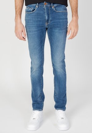 Jeans de mezclilla azul desgastados con corte ajustado, que presentan un cierre de botón y cinco bolsillos. Combinados con zapatillas blancas para un contraste.