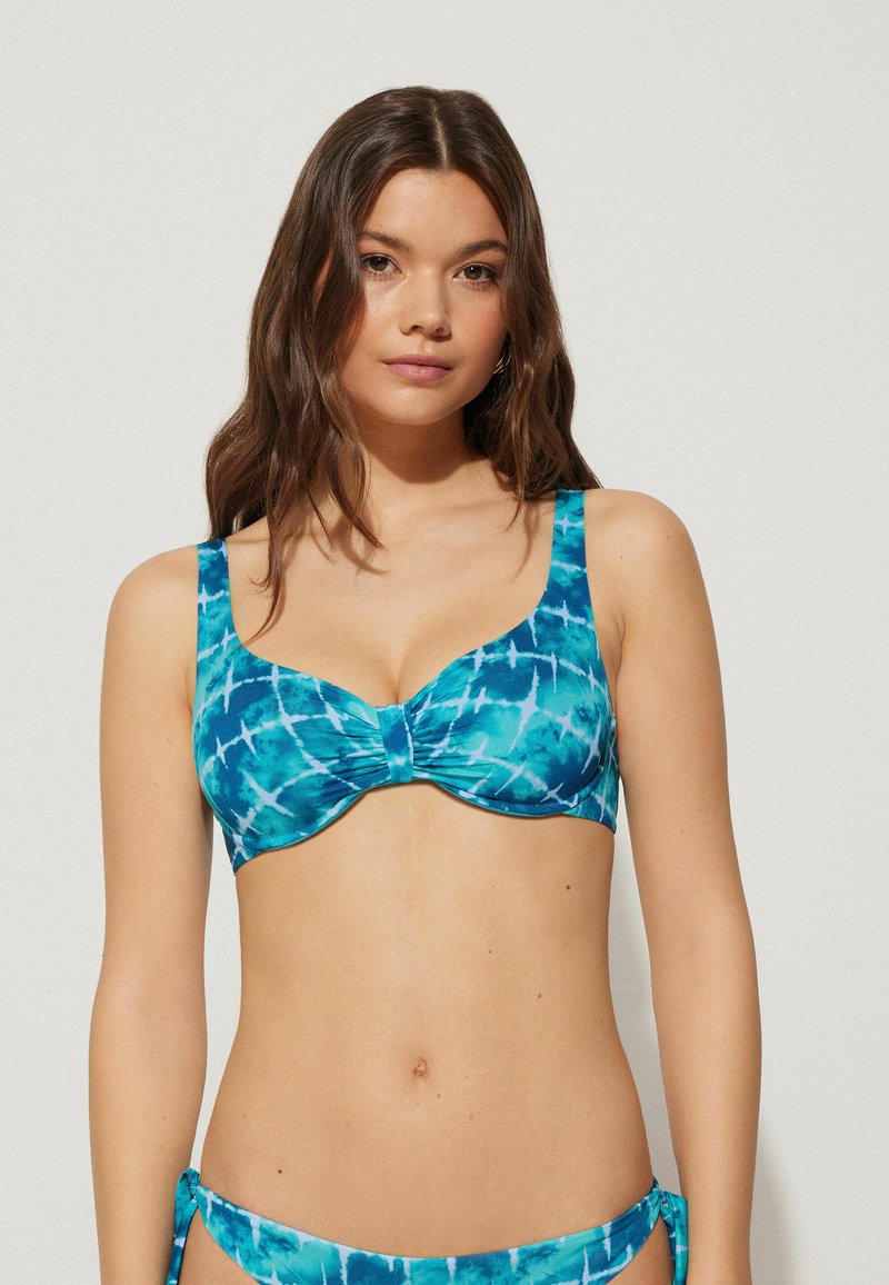 Haut de bikini tie-dye turquoise avec larges bretelles, soutien-gorge avec armatures et détail central froncé. Tissu lisse et extensible avec une finition brillante.