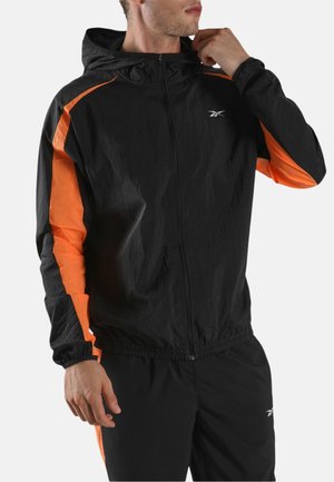 Mann richtet schwarze, mit einem Reißverschluss versehene Kapuzenjacke mit orangefarbenen Streifen an den Ärmeln, kombiniert mit passenden schwarzen Hosen mit orangefarbenen Seitenpaneelen.