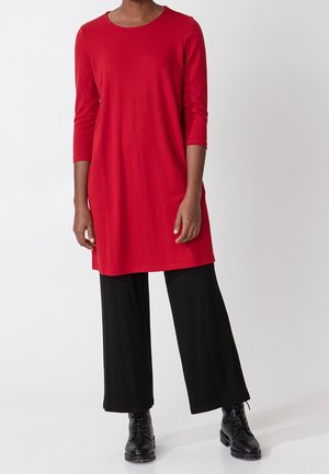 Person iført en lang rød tunika med trekvartærmer, sorte bukser med vide ben og sorte snørestøvler, stående mod en hvid baggrund.