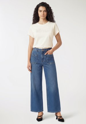 Donna con capelli ricci che indossa una t-shirt bianca con la scritta "Wild Heart", jeans blu a vita alta e gamba larga, e tacchi neri con cinturini, in piedi con una mano in tasca.