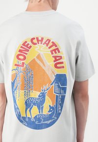 Szürke pamut póló, színes grafikai nyomattal, amelyen szarvas, hegy és ház látható, piros, félkövér betűkkel írva: 'LONE CHATEAU'.
