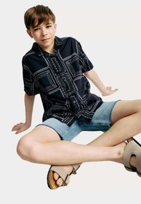 Camisa de manga corta negra con estampado y detalles en blanco, combinada con shorts de mezclilla y sandalias beige claras, posada en una posición sentada.