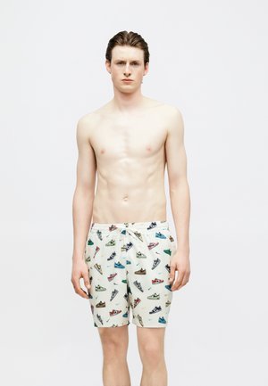 Joven de pie sin camiseta, con pantalones cortos blancos con estampado de zapatillas coloridas, sobre un fondo claro y sencillo.