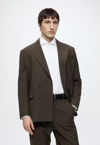 Jeune homme portant un costume croisé marron foncé avec une chemise blanche, debout devant un fond clair uni.