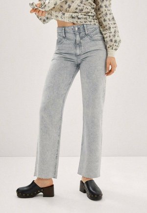 Kobieta ubrana w jasnoszare, wysokie proste jeansy, kwiecistą bluzkę z bufkami na rękawach oraz czarne drewniane chodaki na obcasie z ćwiekami.