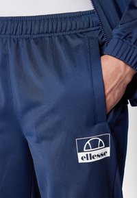 Navy athletic joggers made of smooth fabric, featuring an elastic waistband and side pockets; adorned with a white ellesse logo.