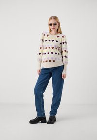 Maglione lavorato a maglia con motivi di cuori colorati in rosa, marrone e giallo su uno sfondo chiaro. Maniche lunghe e a sbuffo; bordi e orlo a coste.