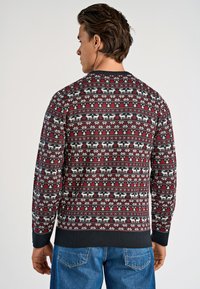Stickad tröja i mörkröd, med ett fair isle-mönster som visar vita renar, träd och geometriska former. Ribbad halsringning och nederkant i mörkblått.