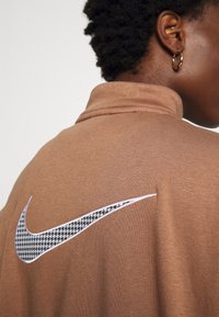Brun sweatshirt med hög krage och en vit Nike swoosh-logotyp, med ett svart hundtandsmönster på baksidan. Mjuk textur, avslappnad passform.
