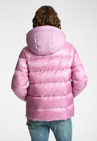 Person som bär en blank pink puffjacka med en stor huva, sedd bakifrån, i kombination med blå denimjeans.