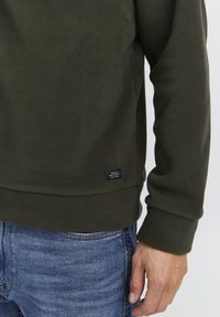Felpa in pile verde scuro con superficie texturizzata, polsini a coste e una piccola etichetta in tessuto all'orlo. Indossata sopra jeans blu.