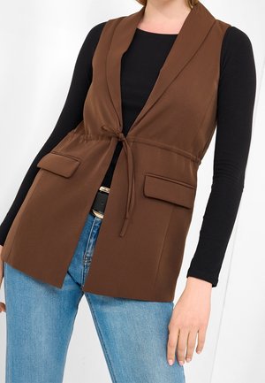 Kobieta w brązowej kamizelce bez rękawów zawiązanej z przodu, na czarnym topie z długim rękawem i jasnoniebieskich dżinsach na białym tle.