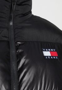 Chaqueta de plumas negra con un collar alto, que presenta una cremallera central y un parche de logo bordado en azul, blanco y rojo en la parte frontal.