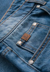 Blå denimtyg med orange sömmar, prydt med metalldragkedjor och en fyrkantig brun läderlapp med en logotyp nära bakfickan.