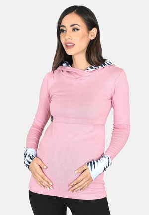 Woman with dark hair wearing a pink long-sleeve top with patterned cuffs, gently holding her pregnant belly, looking to the side.