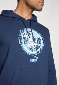 Felpa navy realizzata in morbido tessuto. Presenta una grande grafica del Manchester City in azzurro chiaro con logo centrale e tasca frontale.