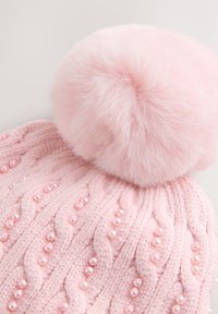 Lyserød strikket hue med en stor imiteret pelspompon. Har et tekstureret kabelstrikket mønster og små dekorative perler over det hele.