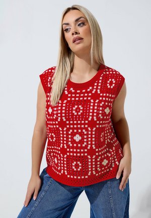 Frau mit langen blonden Haaren, die ein rotes ärmelloses gemustertes Stricktop und blaue Jeans trägt, posiert mit einer Hand auf der Hüfte vor weißem Hintergrund.