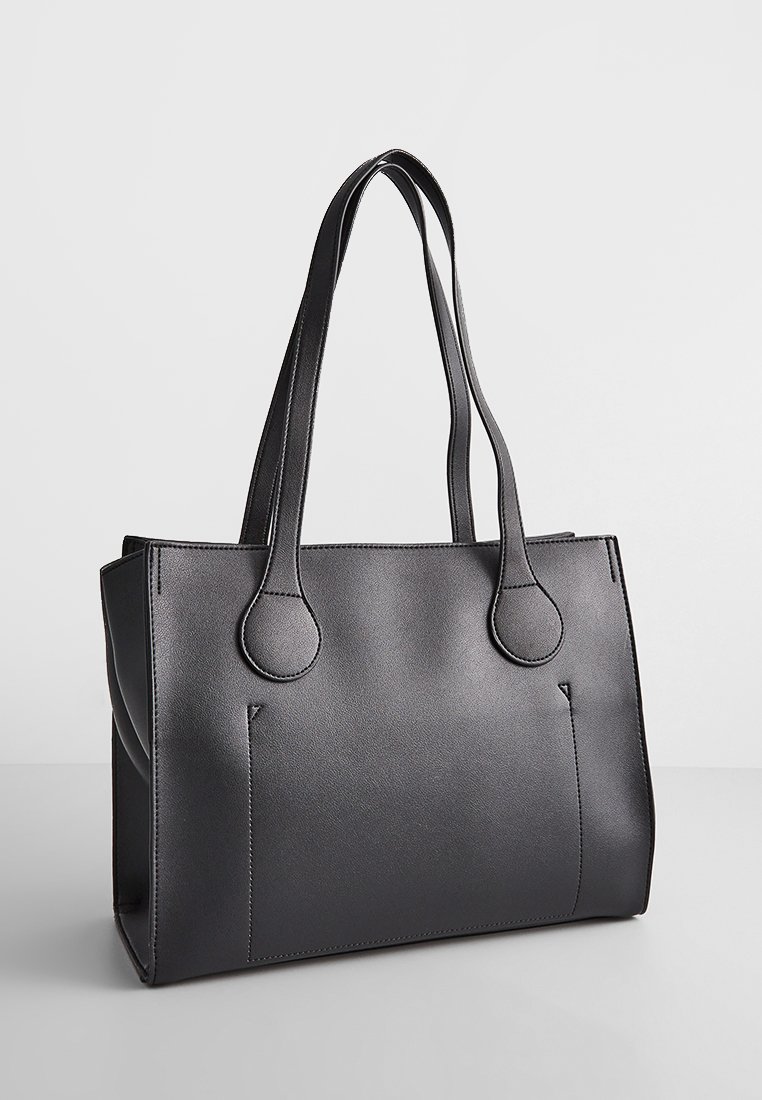 Sac fourre-tout en cuir noir avec poignées doubles, forme structurée et couture visible. Présente deux poches avant et une texture lisse.