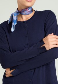 Cardigan blu navy con chiusura a bottoni, maniche a costine con ritagli e una tasca. Indossato con una sciarpa di seta stampata attorno al collo.