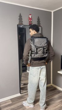 Grå stofrygsæk med justerbare stropper, rullelukning og sorte accenter; placeret foran et spejl med trægulv og grå væg.