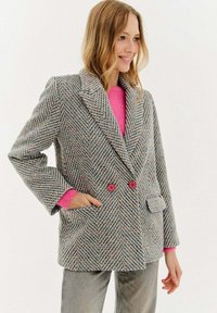 Žena v šedém herringbonovém blejzru s růžovými knoflíky, pod ním má jasně růžový svetr a šedé kalhoty. Usmívá se a pózuje s jednou rukou v kapse.